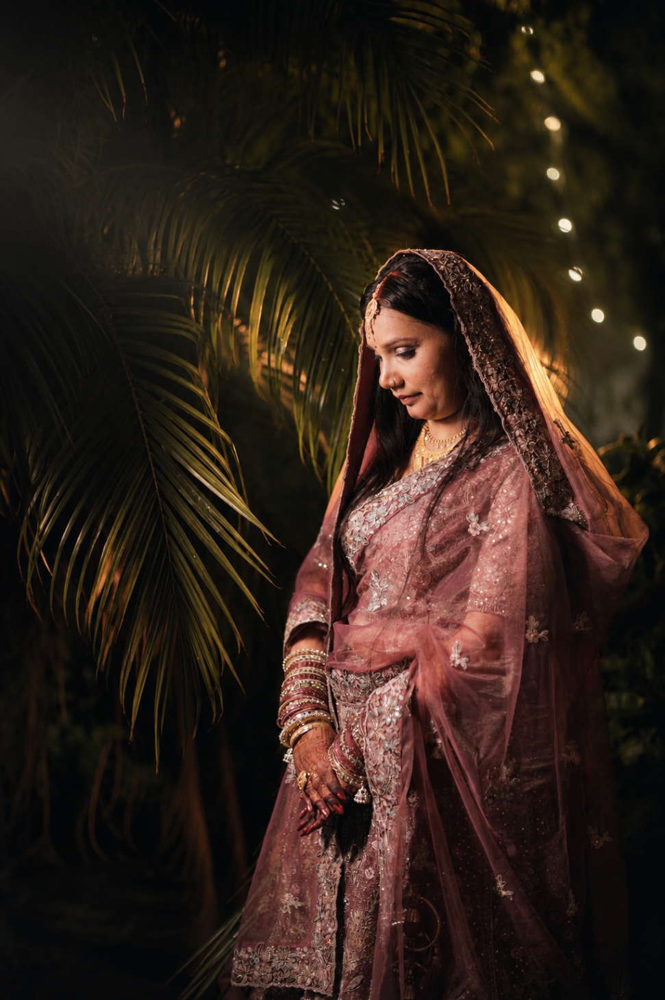 Sakshi in a rose-gold saree standing beneath palm fronds strung with fairy lights