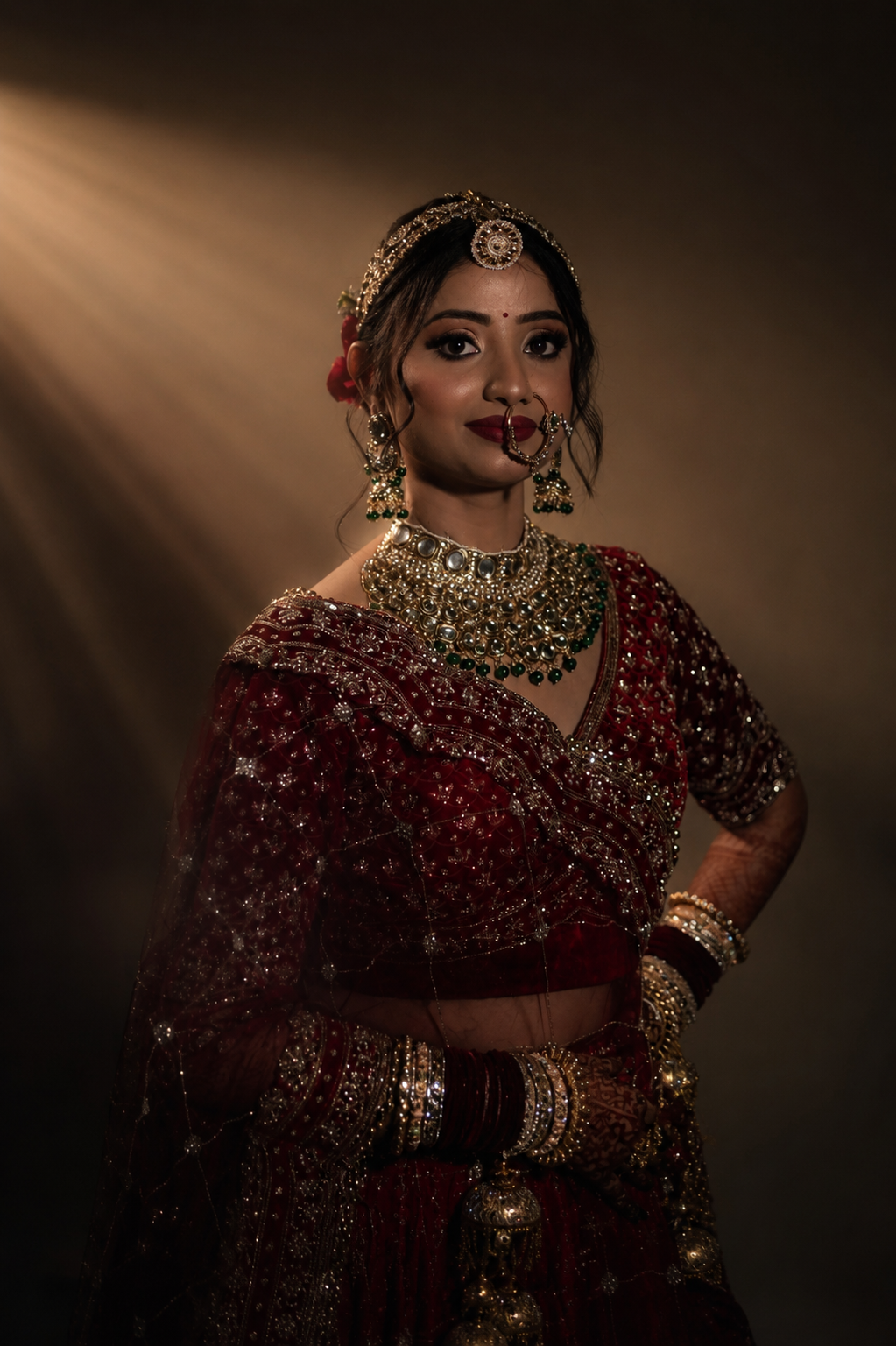 Vaishnavi & Abishek — Vaishnavi in a deep crimson lehenga, studio portrait under a warm shaft of light