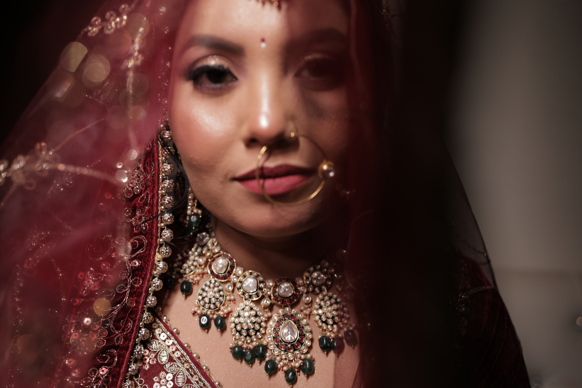 Close portrait of the bride through a crimson veil, kundan necklace catching light