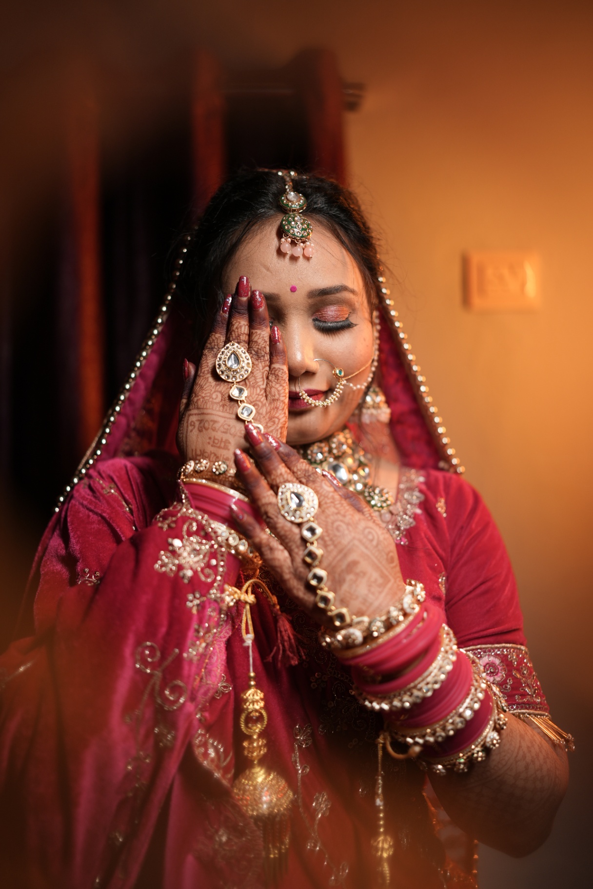 Sakshi in her bridal red, eyes closed, hands framing her hennaed face during a quiet getting-ready moment