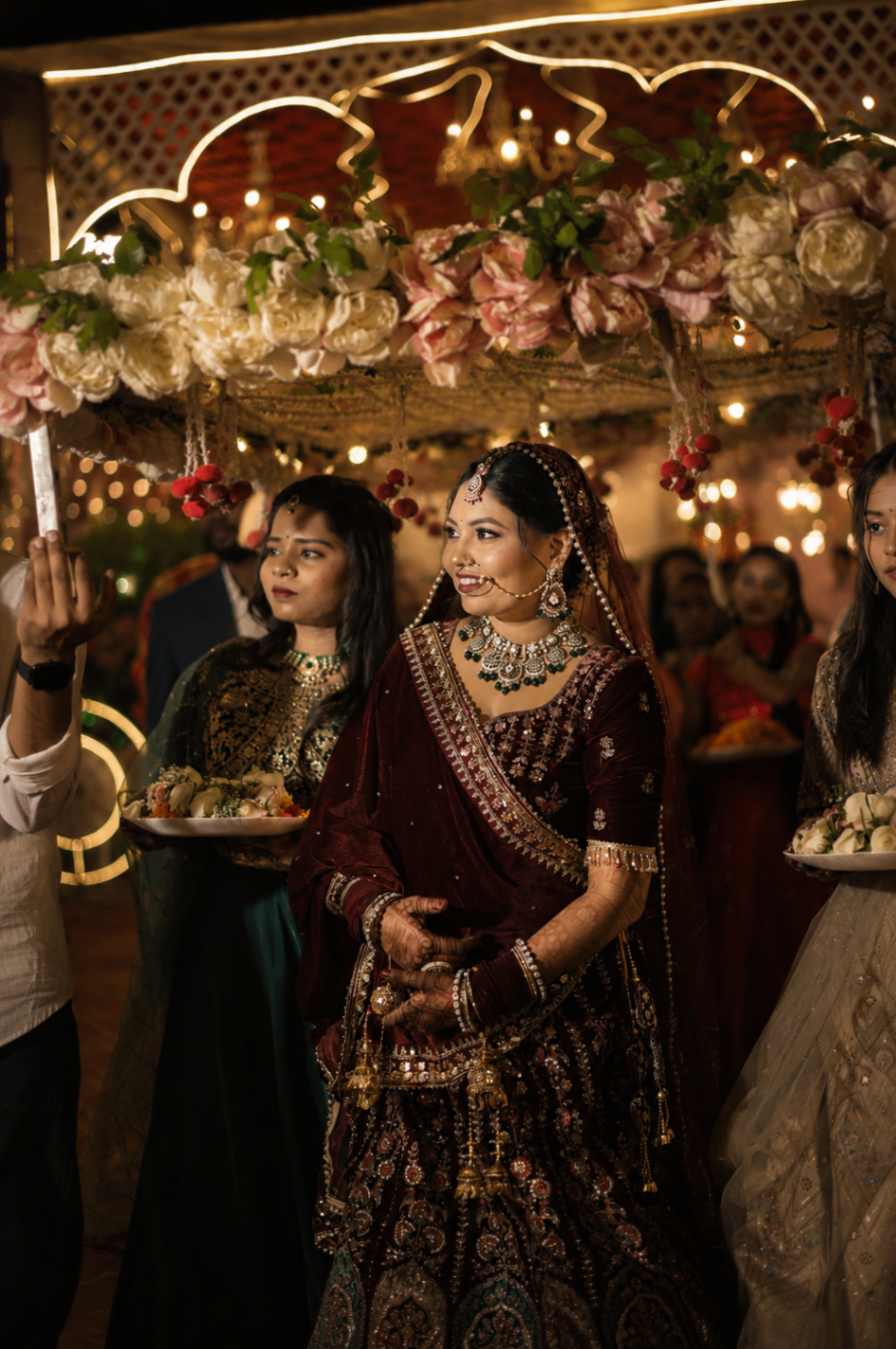 Renu enters beneath a floral canopy of peonies and marigold, deep maroon velvet lehenga catching the fairy lights
