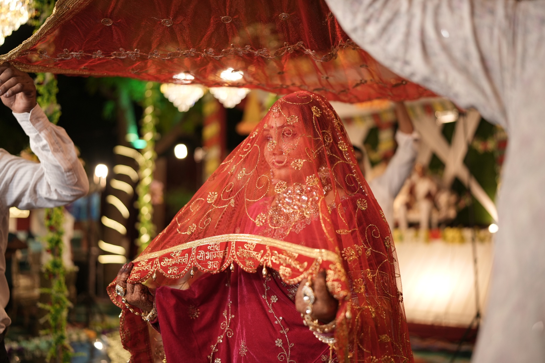 Sakshi walking under a red ceremonial dupatta held aloft by family during the phera ritual