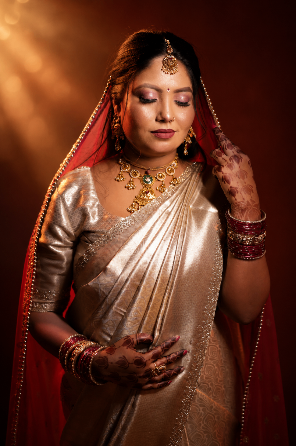 Studio portrait — Renu in a gold silk saree and red veil, eyes lowered as a beam of light crosses her shoulder