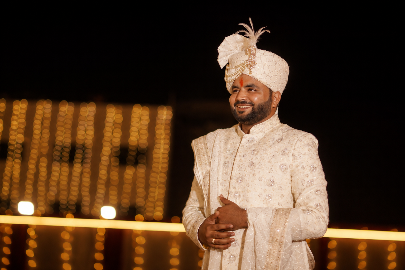 Abishek in ivory sherwani, feathered safa, warm bokeh lights behind him